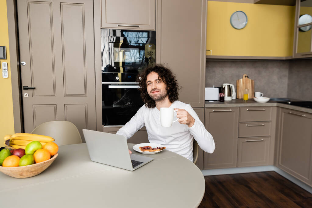 χαμογελαστός ελεύθερος επαγγελματίας κρατώντας φλιτζάνι καφέ και κοιτάζοντας κάμερα κοντά σε φορητό υπολογιστή, προπόσεις και φρέσκα φρούτα - Φωτογραφία, εικόνα