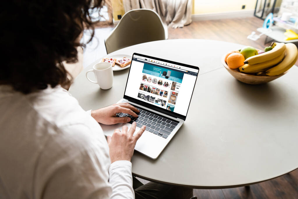 KYIV, UKRAINE -エイプリル社202020年25日:アマゾンウェブサイトのノートパソコンを使って、果物、コーヒーカップ、トースト付きのプレートの近くでフリーランスのバックビュー - 写真・画像