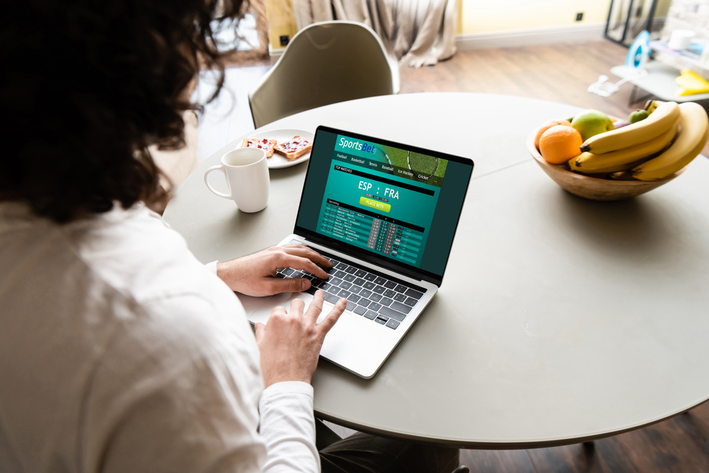 πίσω όψη του freelancer χρησιμοποιώντας φορητό υπολογιστή με την ιστοσελίδα Sportsbet κοντά σε φρούτα, φλιτζάνι καφέ και πιάτο με προπόσεις - Φωτογραφία, εικόνα