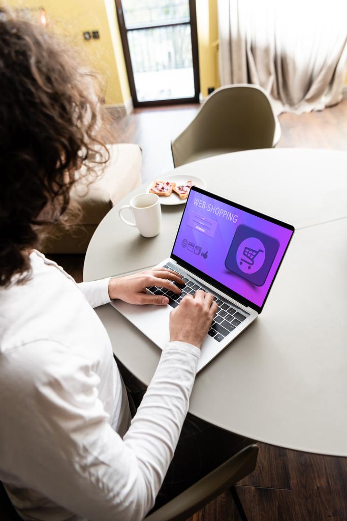 vista de ángulo alto del freelancer utilizando el ordenador portátil con el sitio web de compras web cerca de taza de café y plato con tostadas
 - Foto, Imagen