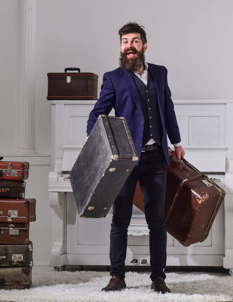 Man, traveller with beard and mustache with luggage, luxury white interior background. Macho elegant on smiling face stands near pile of vintage suitcase, holds suitcases. Baggage delivery concept. - Photo, Image
