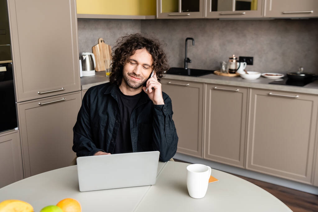 Lächelnder Freiberufler, der in der Küche neben der Kaffeetasse mit dem Laptop auf dem Smartphone spricht - Foto, Bild