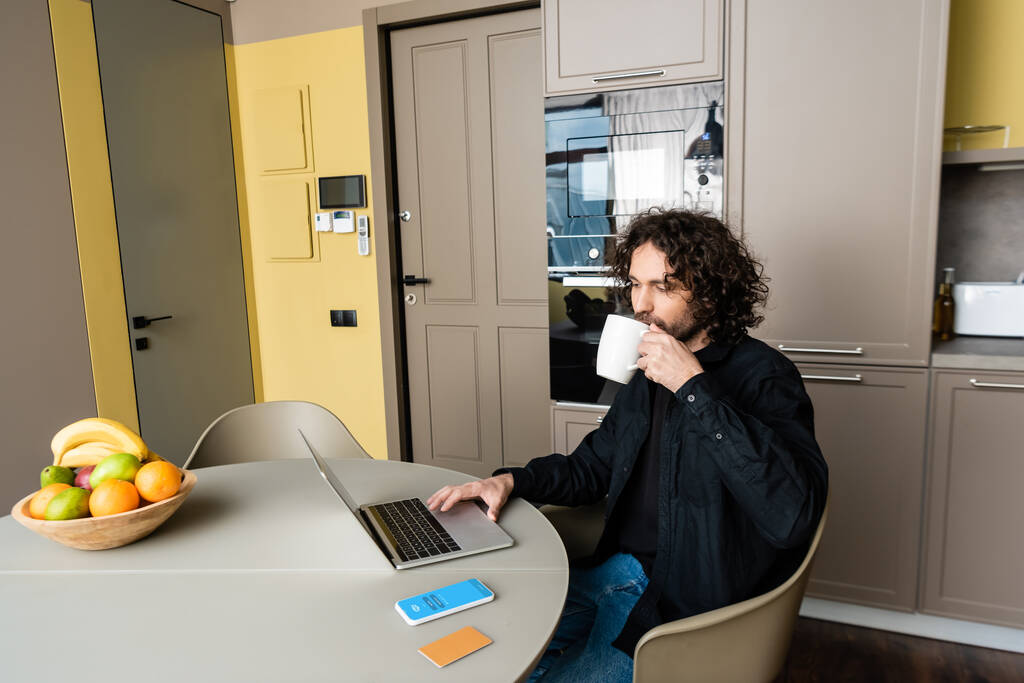 KYIV, UKRAINE - 25 de abril de 2020: belo freelancer bebendo café enquanto usa laptop perto do smartphone com aplicativo Skype, cartão de crédito e frutas frescas
 - Foto, Imagem