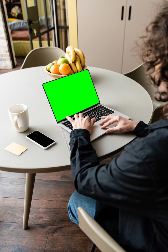 achteraanzicht van freelancer met behulp van laptop met groen scherm in de buurt van smartphone, koffiebeker en kom met vers fruit - Foto, afbeelding