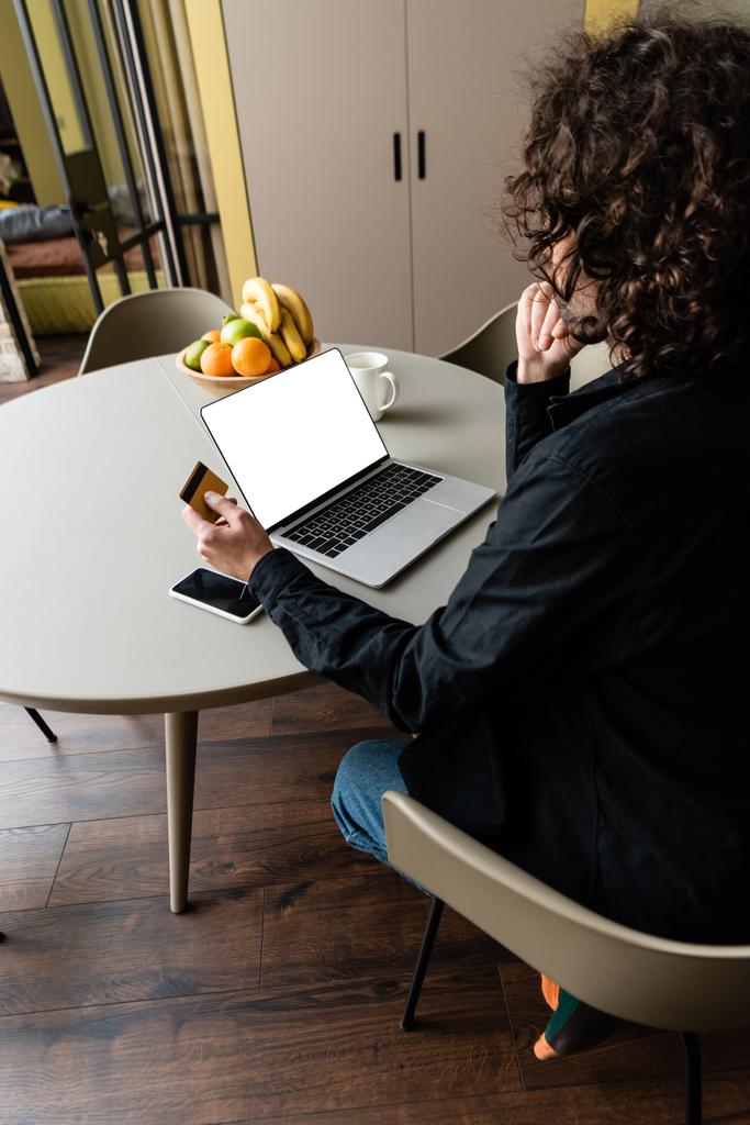 back view of freelancer holding credit card while sitting at laptop with white screen, smartphone and fruits - Photo, Image