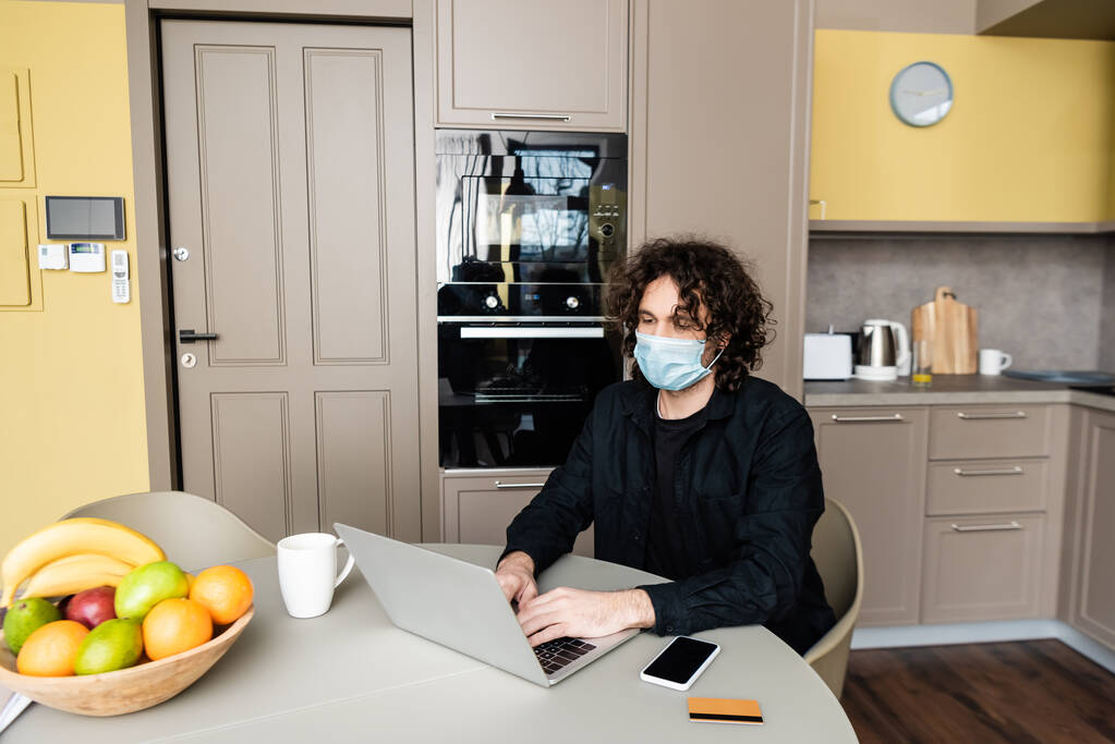 スマートフォンの近くにノートパソコンを使用して医療用マスクの男,キッチンテーブルの上のクレジットカードや果物  - 写真・画像