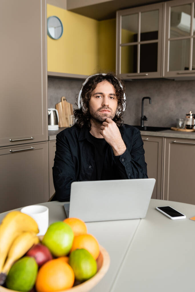 Kulaklıklı bir adamın bilgisayarın yanındaki kameraya, akıllı telefona ve mutfaktaki masada duran meyvelere seçici bir şekilde bakması.  - Fotoğraf, Görsel