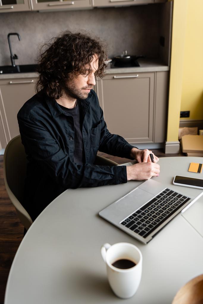 Селективное внимание человека, пишущего рядом с цифровыми устройствами и кредитной картой во время вебинара на кухне
   - Фото, изображение