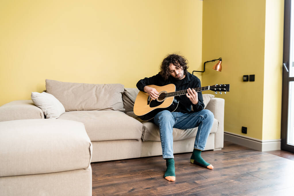 Homme bouclé jouant de la guitare acoustique assis sur le canapé à la maison
  - Photo, image