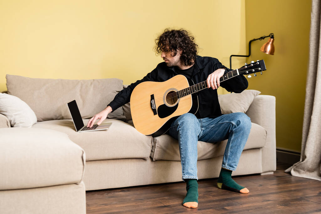 Curly férfi gazdaság akusztikus gitár és a laptop használata az online oktatás során a nappaliban - Fotó, kép
