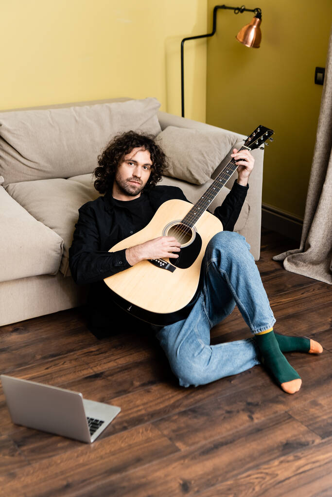 Komea mies pitelee akustista kitaraa ja katsoo kameraa läppärin lähellä lattialla
  - Valokuva, kuva