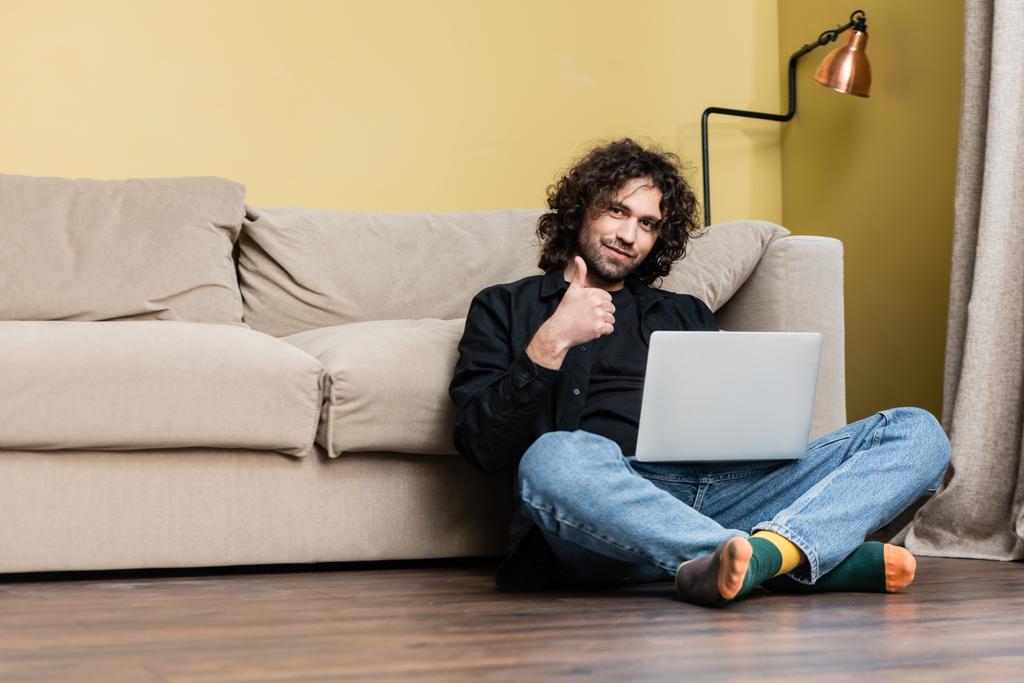 Visão de baixo ângulo de homem bonito mostrando o polegar para cima enquanto trabalhava no laptop no chão
  - Foto, Imagem