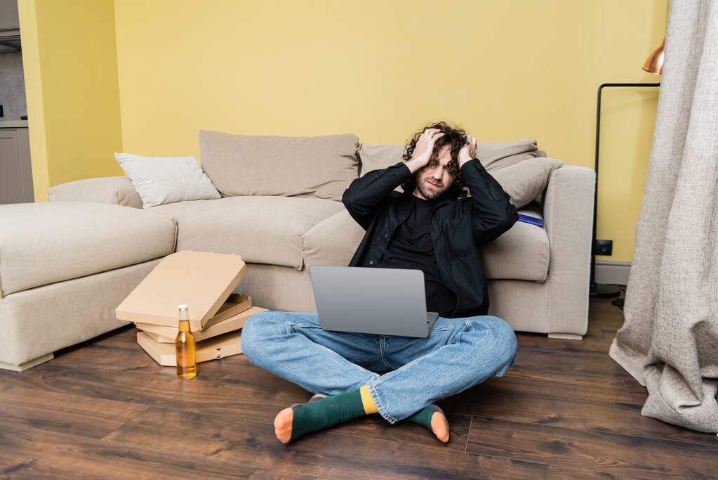 Uitgeputte man die laptop vasthoudt terwijl hij op de vloer zit in de buurt van bierflesjes en pizzadozen  - Foto, afbeelding