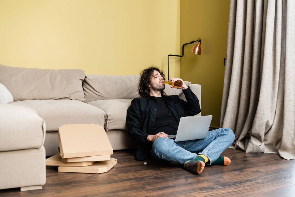 Enfoque selectivo del hombre bebiendo cerveza mientras usa el portátil cerca de cajas de pizza en el suelo
  - Foto, Imagen