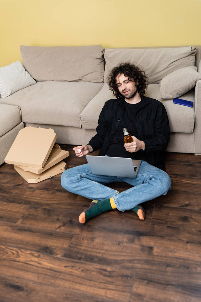 Curly teleworker holding bottle of beer and using laptop near pizza boxes in living room - Photo, Image