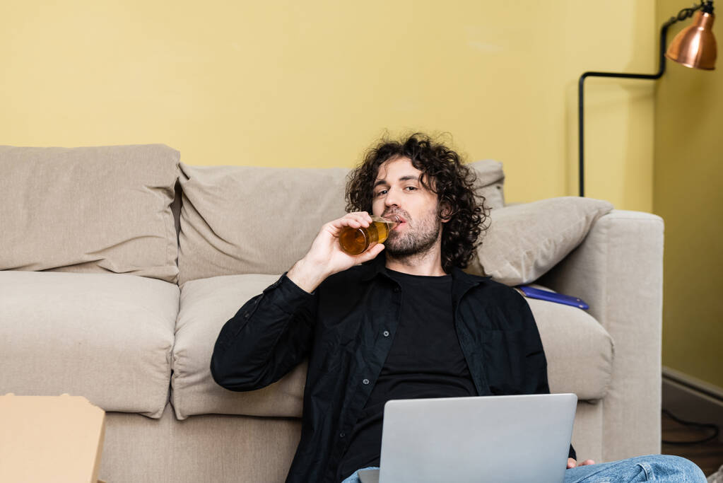 Όμορφος άνθρωπος κοιτάζοντας κάμερα, ενώ πίνοντας μπύρα κοντά στο laptop και πίτσα κουτί στο σπίτι  - Φωτογραφία, εικόνα