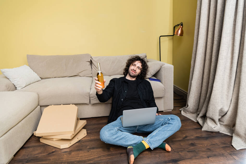 Ο κατσαρομάλλης κοιτάζει την κάμερα κρατώντας ένα μπουκάλι μπύρα κοντά στο λάπτοπ και κουτιά πίτσας στο πάτωμα στο σπίτι  - Φωτογραφία, εικόνα