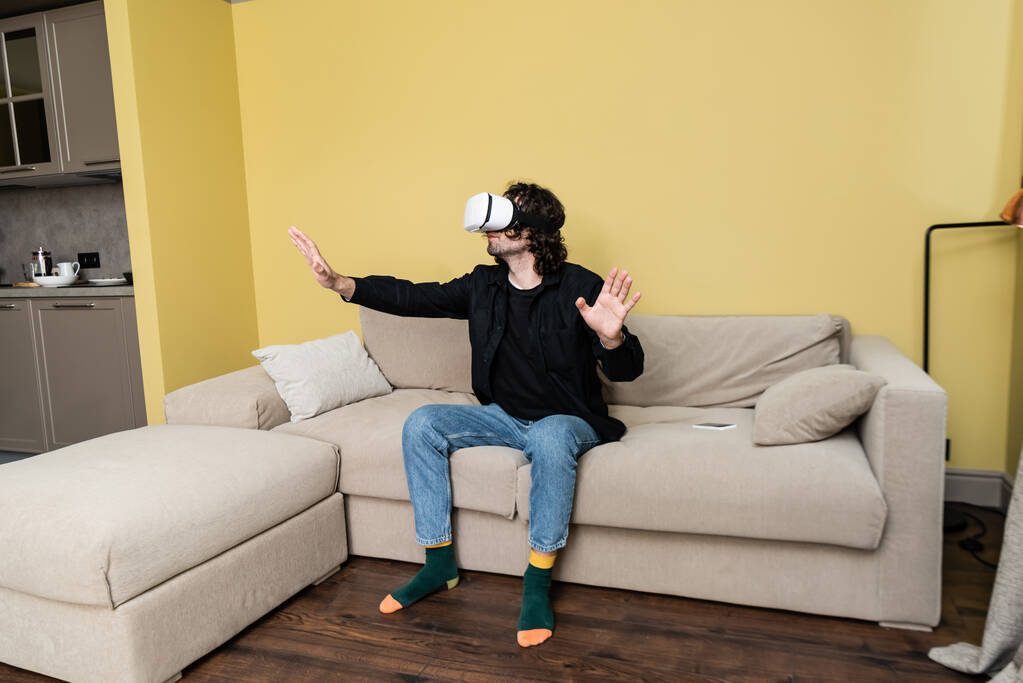 krullende man met behulp van virtual reality headset in de buurt van smartphone op de bank thuis - Foto, afbeelding