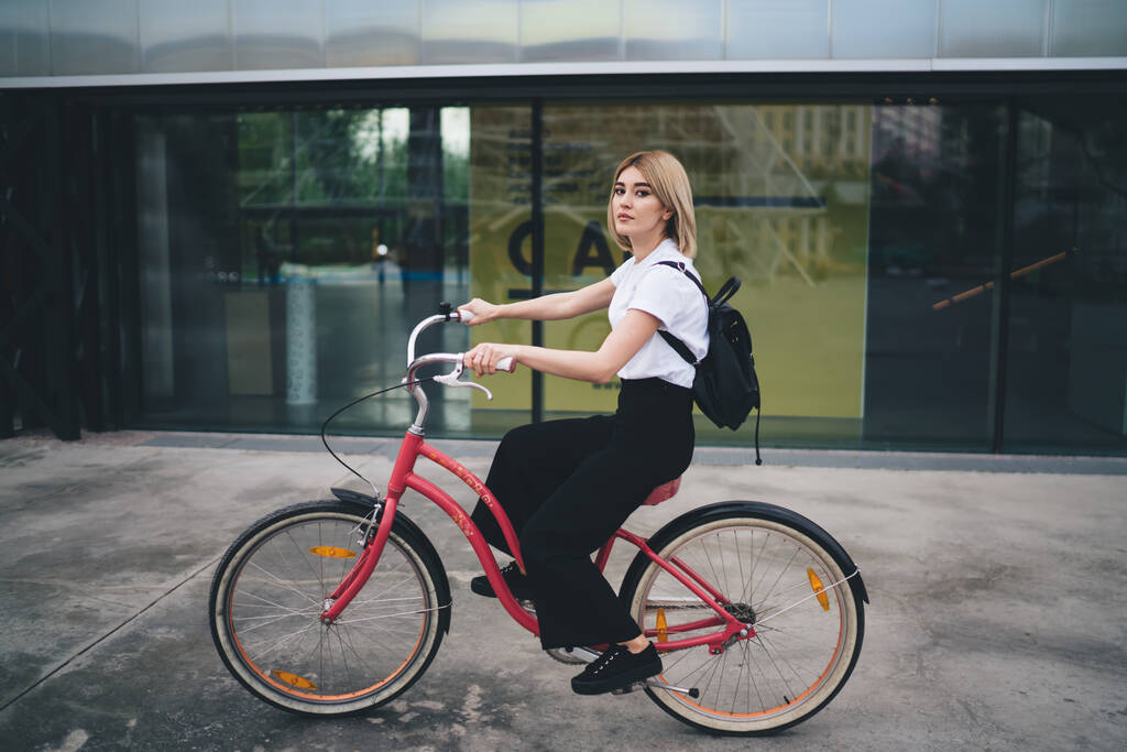Вид сбоку уверенной современной женщины в повседневной одежде и с черным рюкзаком на велосипеде на фоне уличного здания, глядя в камеру - Фото, изображение