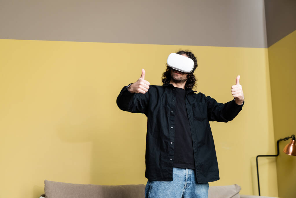 Lockiger Mann mit Kopfhörer zeigt Daumen nach oben im Wohnzimmer - Foto, Bild
