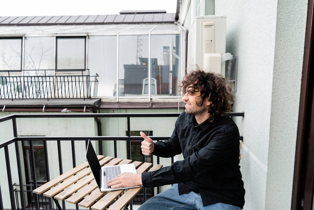 Freelancer sorridente mostrando como gesto ao usar laptop na varanda em casa
  - Foto, Imagem