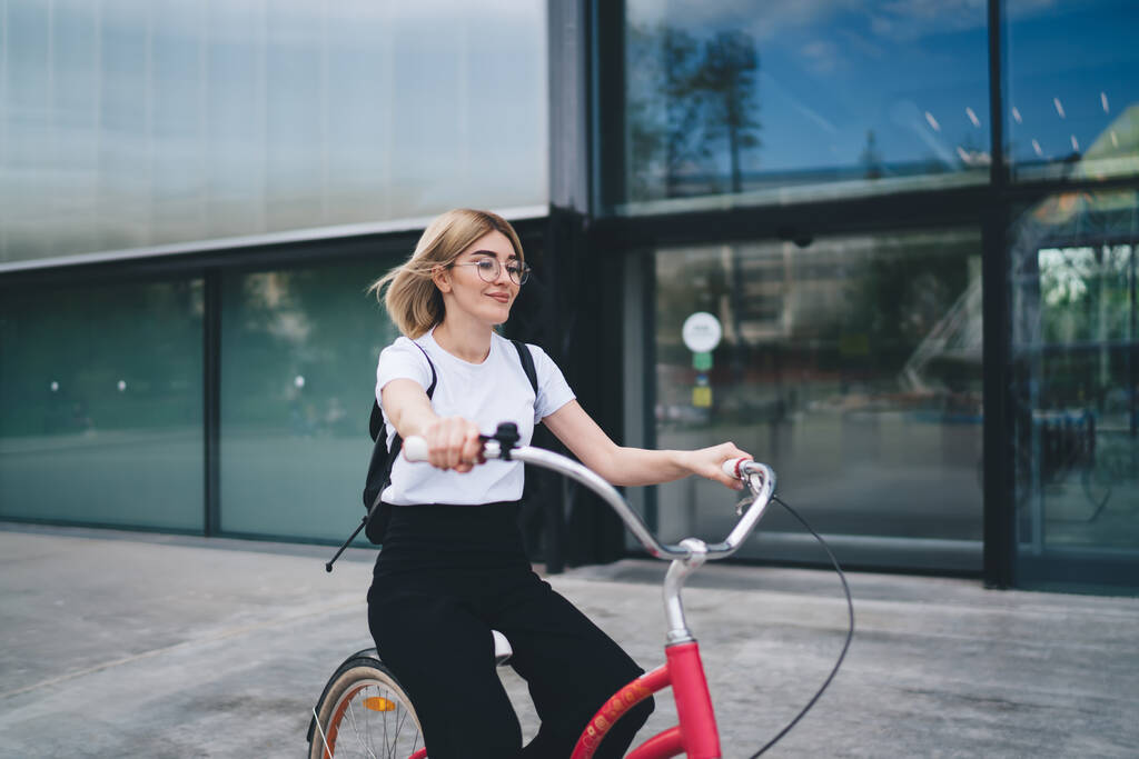 Young pretty woman with backpack dressed in casual clothes and glasses sitting on bicycle saddle holding handlebar while riding on street - Photo, Image