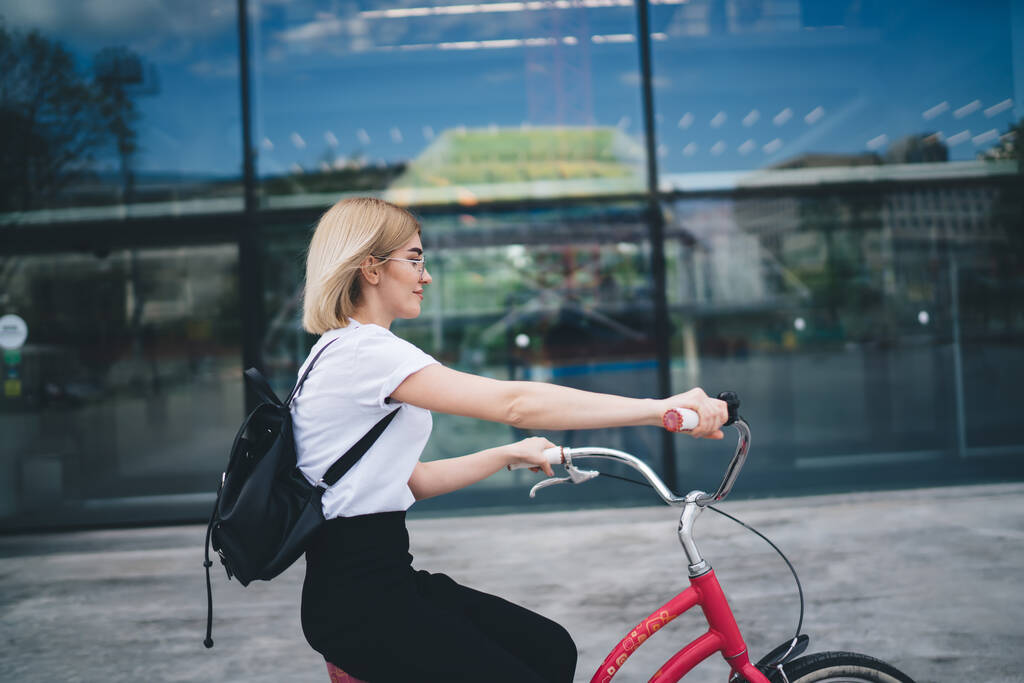 Vue latérale de jeune jolie femme blonde en vêtements décontractés avec sac à dos noir assis sur le vélo et à cheval sur la rue - Photo, image