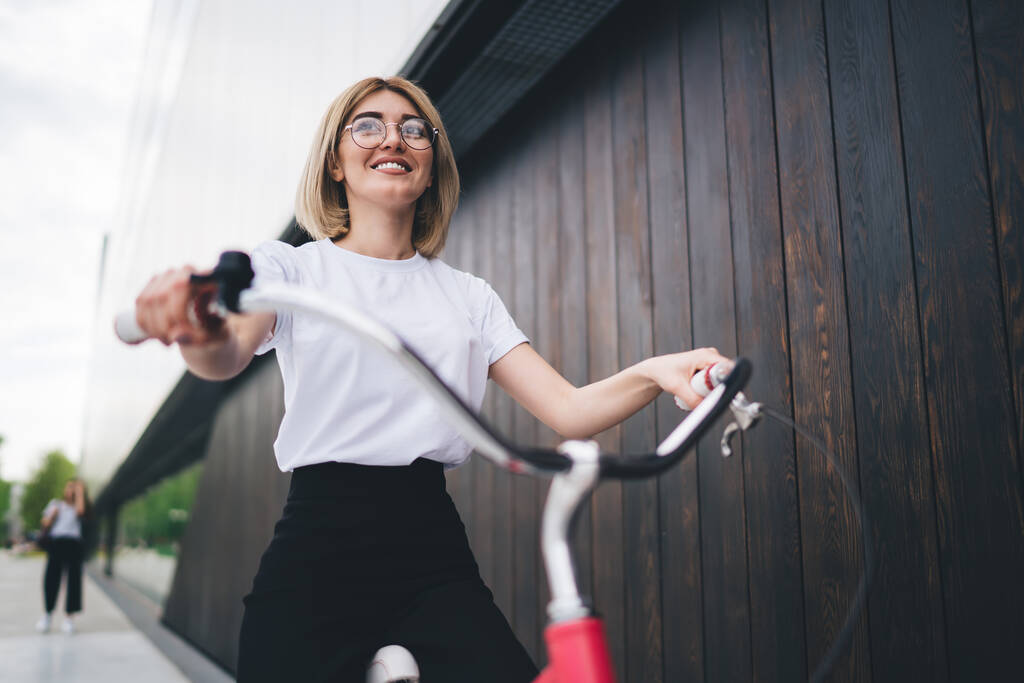 Lage hoek van glimlachende duizendjarige vrouw in terwijl t-shirt en zwarte broek rijden rode fiets langs donker hout plank muur, terwijl kijken vooruit - Foto, afbeelding