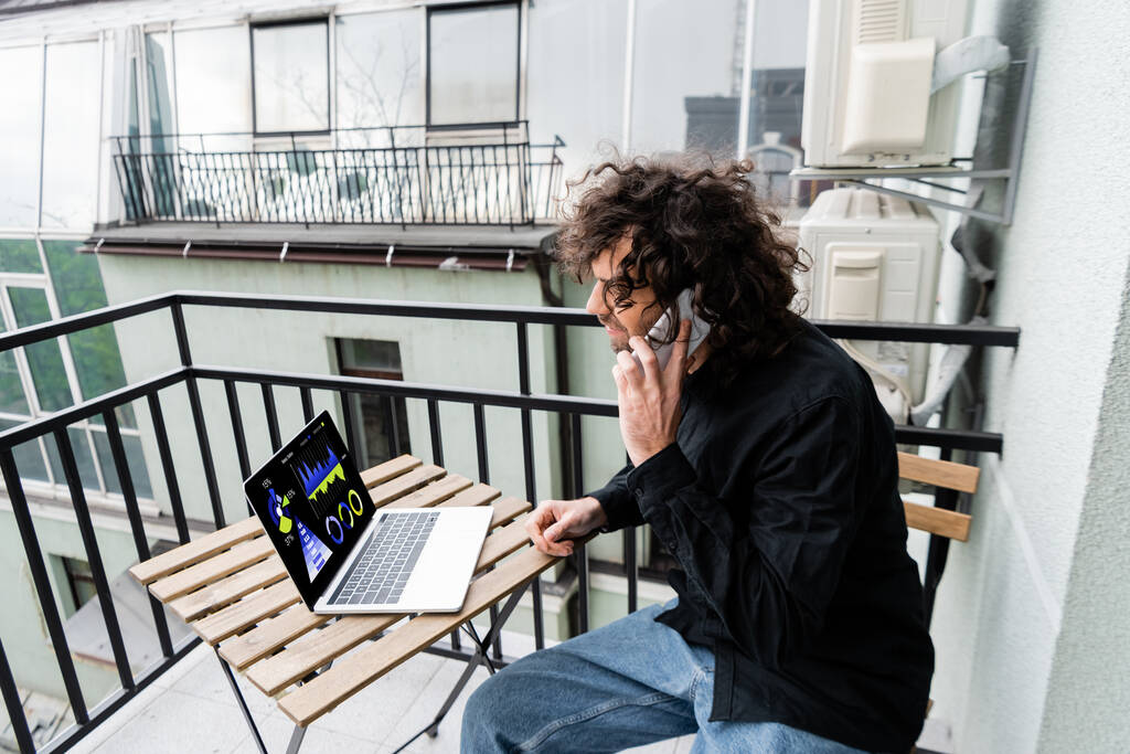 Чоловік розмовляє на смартфоні біля ноутбука з графіками на столі на балконі
  - Фото, зображення