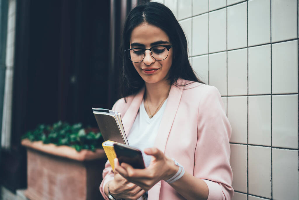 Moderne geschäftige Frau mit Brille und Blazer liest dringende E-Mails ihres Kunden auf dem Smartphone, während sie mit Papieren und Tagebuch in der Hand an der Hauswand steht  - Foto, Bild