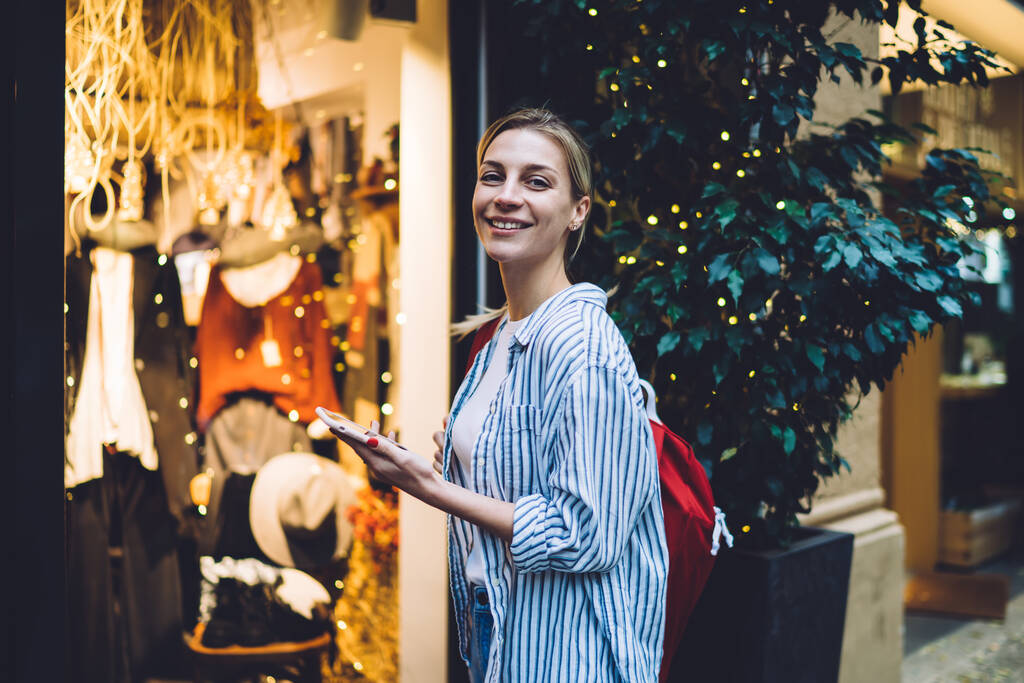 Femme blonde joyeuse en chemise rayée debout et regardant la caméra tout en utilisant un smartphone près du magasin de vêtements sur la rue de la ville - Photo, image