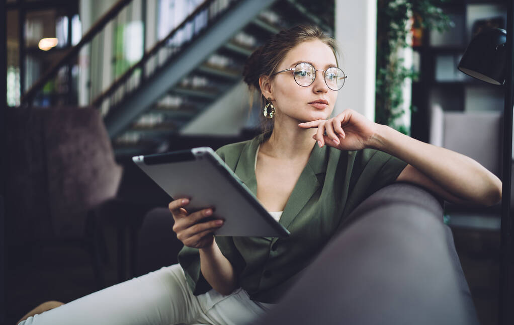Προσεκτική νεαρή γυναίκα σε στρογγυλά γυαλιά κάθεται στον καναπέ αγγίζοντας το πηγούνι και κοιτάζοντας μακριά σε σκέψεις, ενώ κάθεται με tablet στον καναπέ στο γραφείο - Φωτογραφία, εικόνα