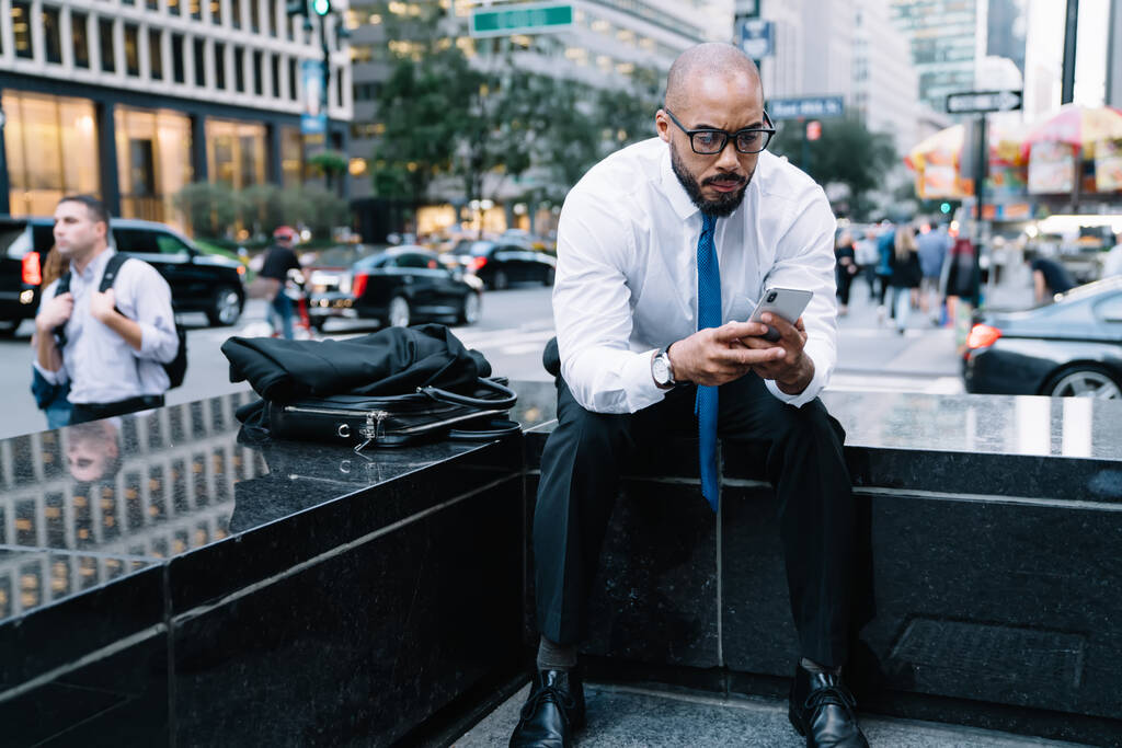 Видатний афроамериканець у сорочці та окулярах читає смартфон, сидячи на вулиці зі шкіряним мішком на задньому плані розмитих будинків та натовпу людей.  - Фото, зображення