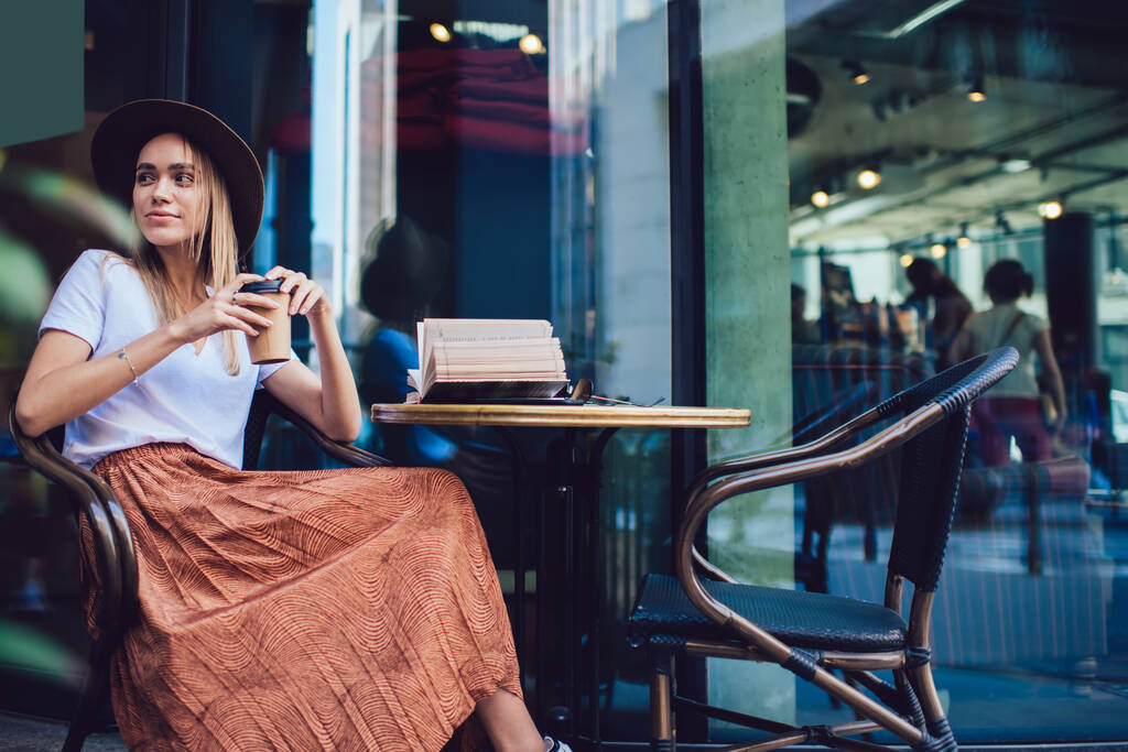Zijaanzicht van zorgeloze mooie jonge vrouw in stijlvolle hoed houden afhaalbeker terwijl zitten aan tafel met geopende boek op coffeeshop straat terras - Foto, afbeelding