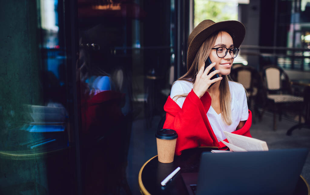 Szczęśliwa uśmiechnięta kobieta w modnej czapce i okularach pokryta kolorowym czerwonym kocem mówiącym smartfonem siedząc przy stole z laptopem i filiżanką kawy w ulicznej kawiarni - Zdjęcie, obraz