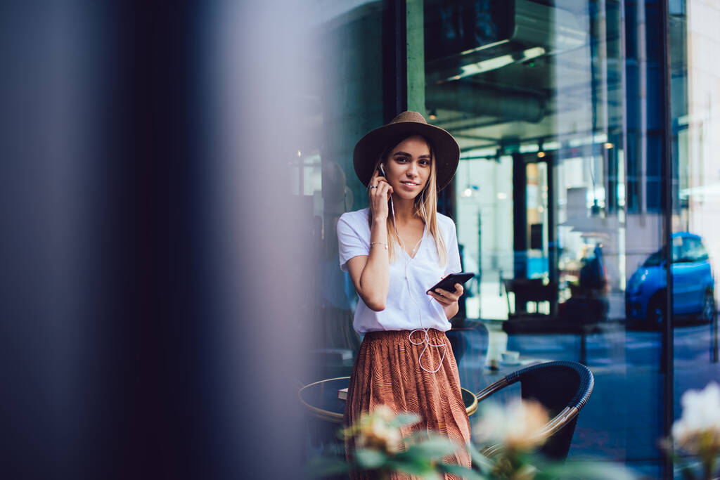 Jeune femme élégante attrayante dans des vêtements à la mode et chapeau avec écouteurs portant smartphone à la main et regardant la caméra tout en se tenant dans un café de rue  - Photo, image