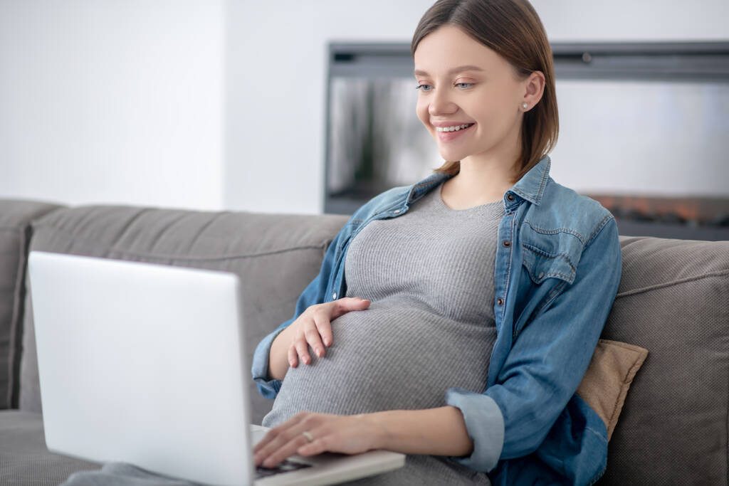 Junge schwangere Frau in Jeansjacke surft im Internet und lächelt - Foto, Bild