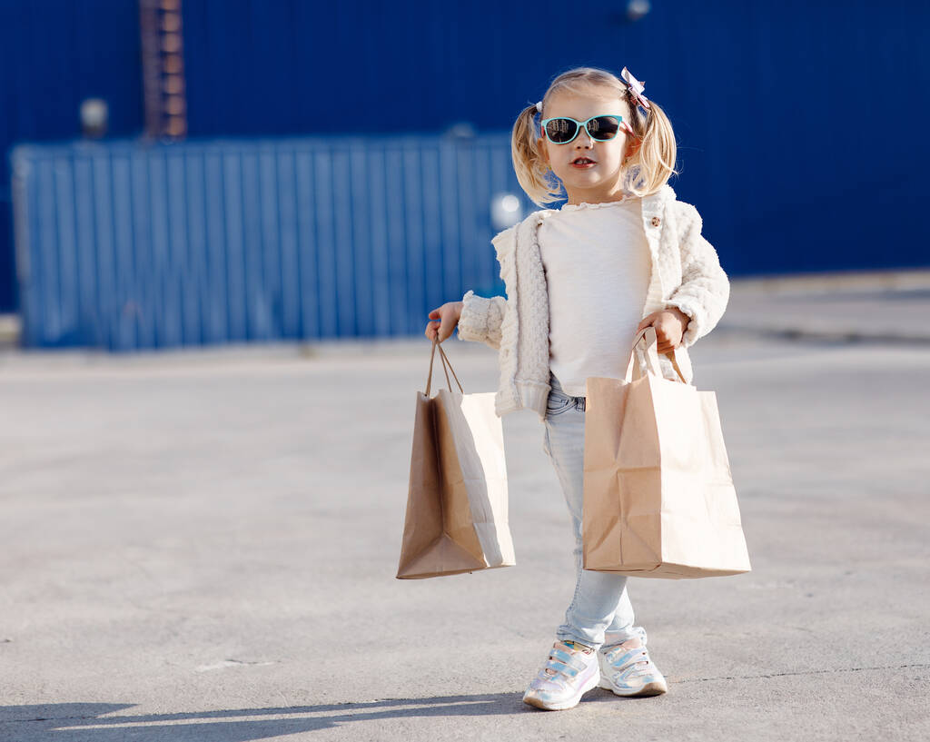 Wesołe dziecko z zakupami. Mała dziewczynka trzyma torby na zakupy.Mała dziewczynka sama na wiosnę na zewnątrz z papierowymi torbami na zakupy.Pretty girl, blondynka w okularach przeciwsłonecznych pozowanie samotnie na ulicy z torbami na zakupy - Zdjęcie, obraz