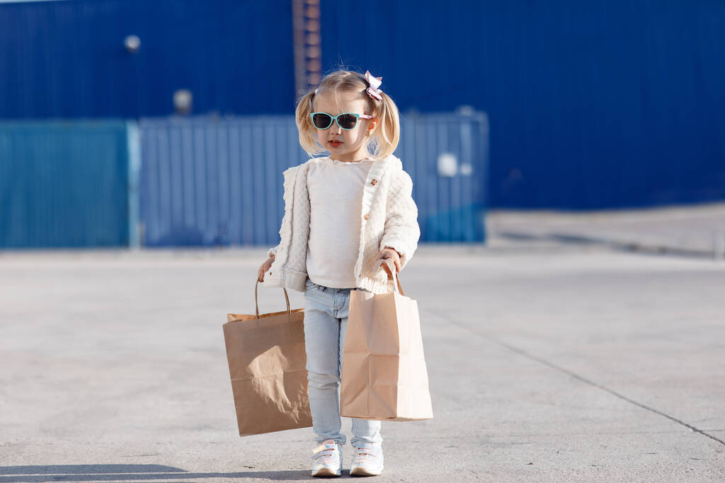 Fröhliches Baby mit Einkäufen. Kleines Mädchen mit Einkaufstaschen.Kleines Mädchen allein im Frühling im Freien mit Papiertüten zum Einkaufen.Hübsches Mädchen, Blondine in Sonnenbrille posiert allein auf der Straße mit Einkaufstaschen - Foto, Bild