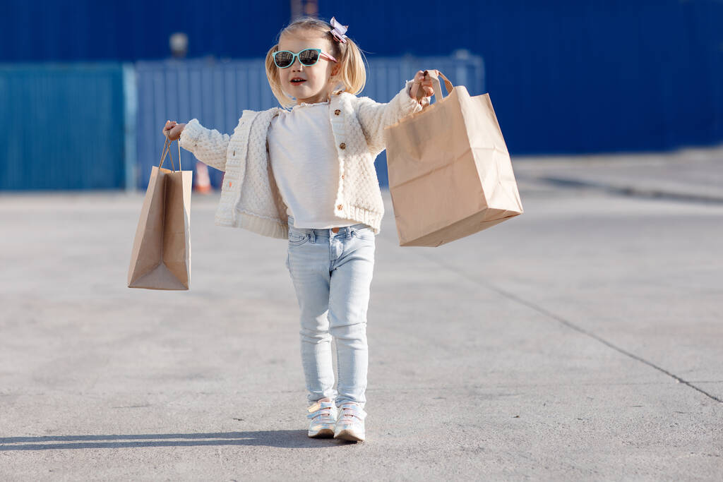 Alımlı neşeli bir bebek. Alışveriş torbaları tutan küçük bir kız. Baharda alışveriş torbalarıyla yalnız başına. Güneş gözlüklü güzel bir kız, alışveriş torbalarıyla sokakta yalnız poz veriyor. - Fotoğraf, Görsel