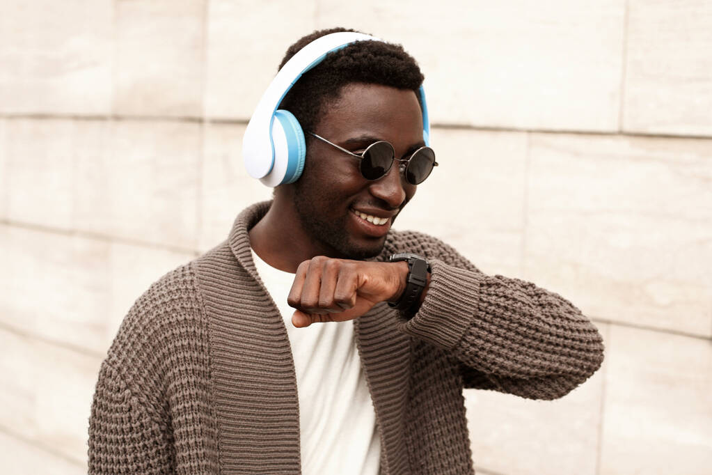 Retrato sorrindo homem africano com smartwatch usando gravador de comando de voz ou leva chamando fones de ouvido sem fio ouvindo música na rua da cidade sobre o fundo da parede de tijolo
 - Foto, Imagem