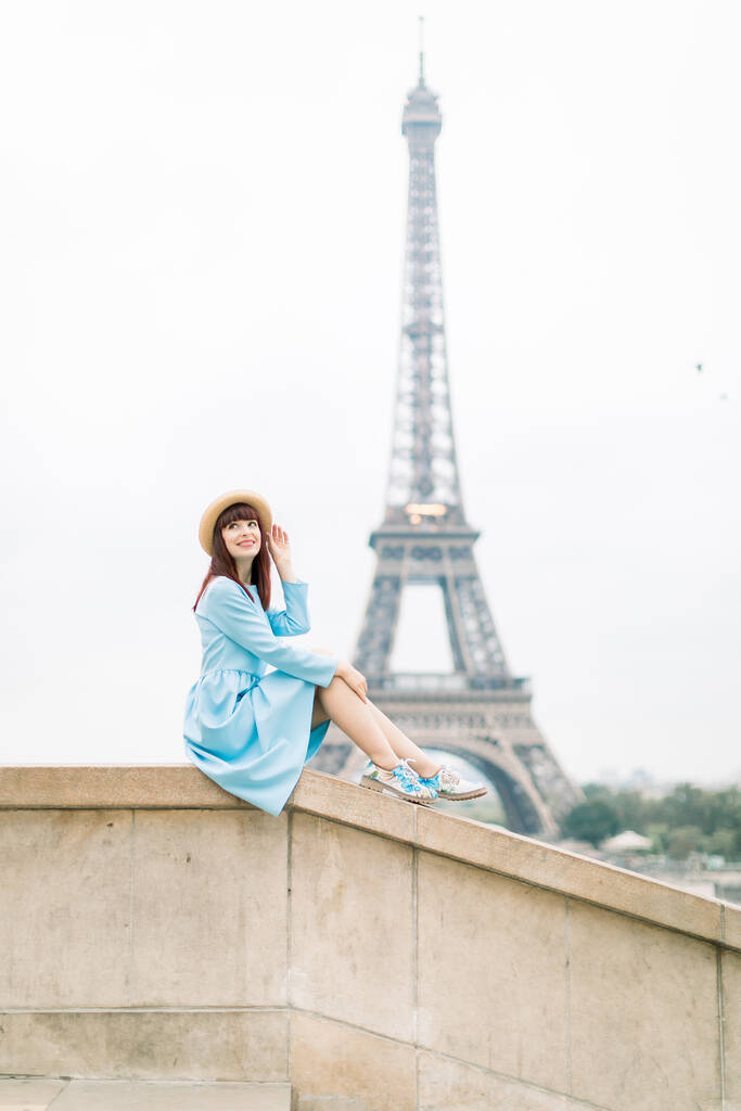 Gyönyörű párizsi vörös hajú lány elegáns kalap és kék ruha ül a lépcsőn az Eiffel torony háttér, és felnézett szép mosollyal - Fotó, kép
