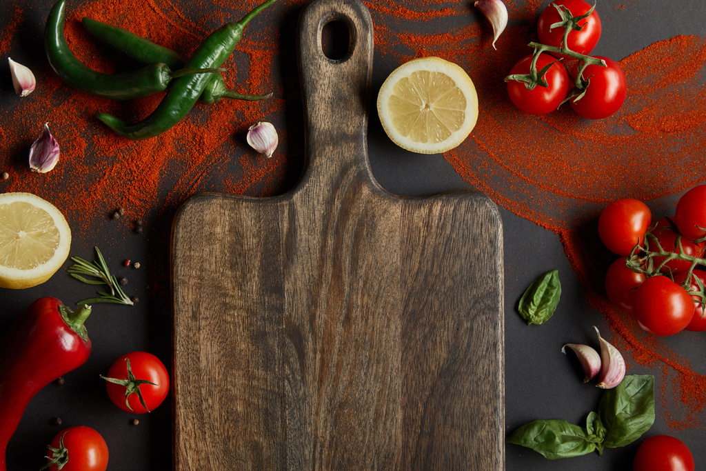 top view of cherry tomatoes, garlic cloves, lemons, green chili peppers, paprika powder and basil leaves near wooden cutting board on black - Photo, Image