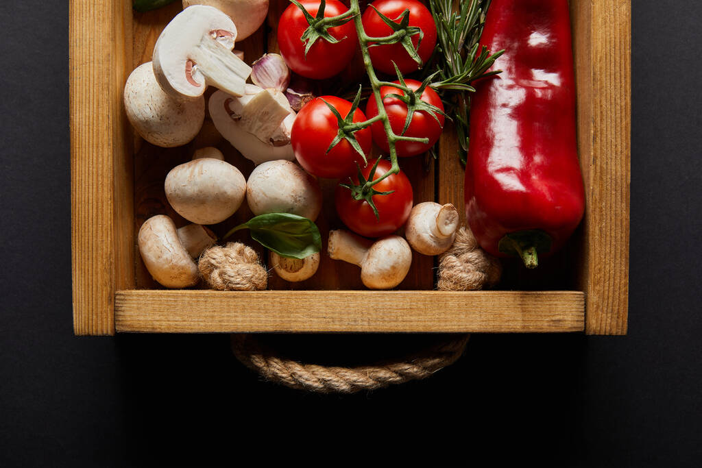 bovenaanzicht van kerstomaten, chili peper, champignons, bloemkool en knoflookteentjes bij basilicum blad en rozemarijn in houten doos op zwart - Foto, afbeelding