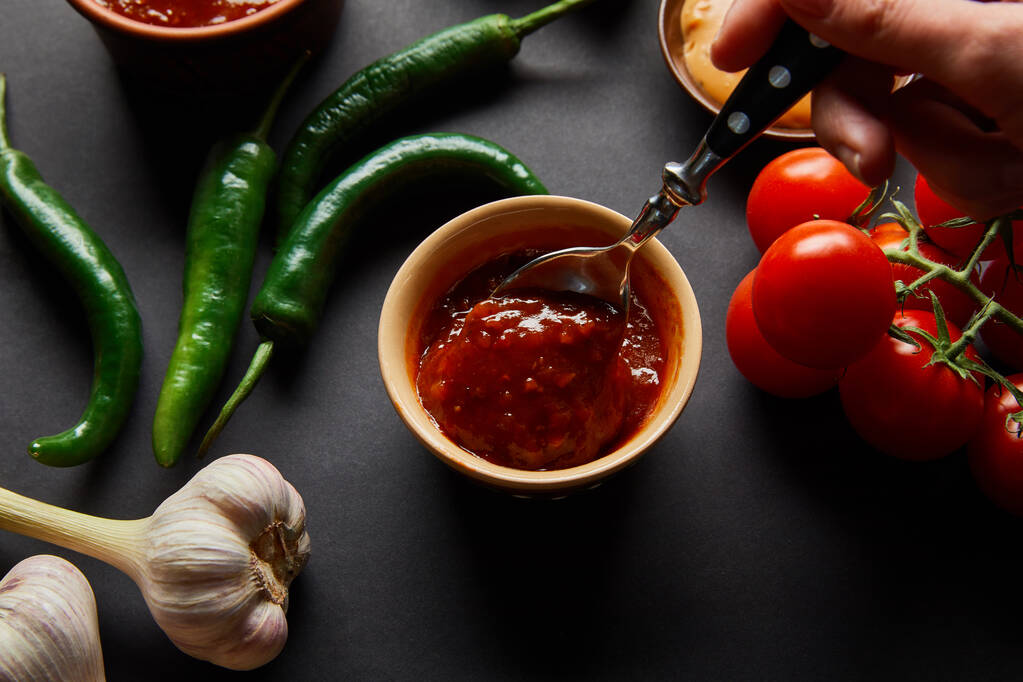 vista recortada de una persona sosteniendo cuchara cerca de sabrosa salsa de tomate y verduras en negro
 - Foto, imagen