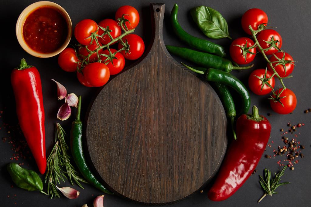 top view of chopping board near ripe cherry tomatoes, garlic cloves, rosemary, peppercorns, basil leaves and green chili peppers on black - Photo, Image