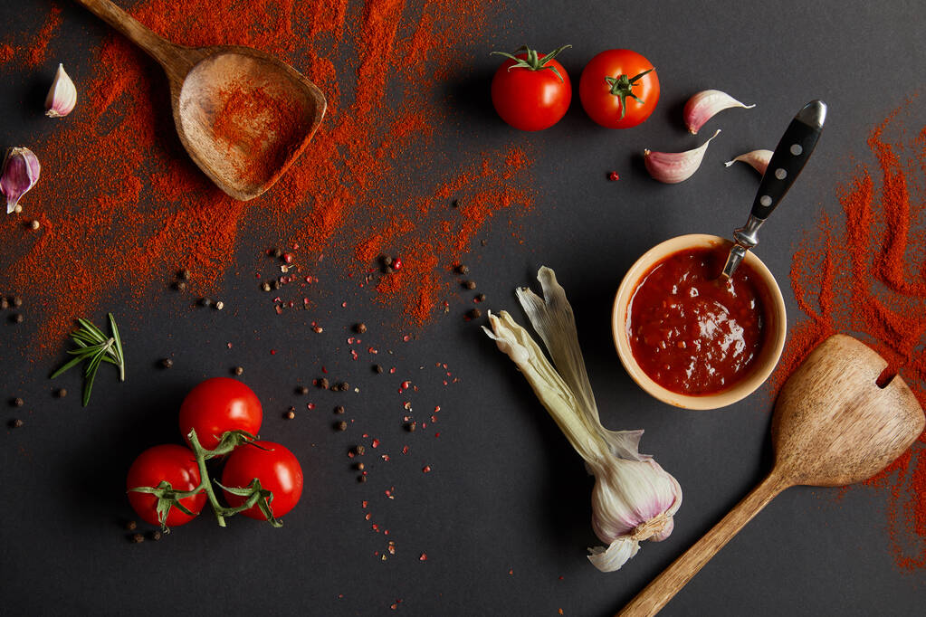 vista superior de tomates cherry maduros, dientes de ajo y hierbas frescas cerca de cucharas de madera con pimentón en polvo sobre negro
  - Foto, imagen