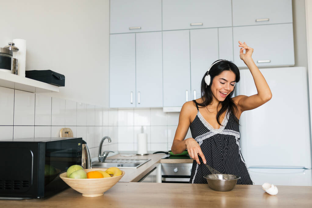 atractiva joven flaca sonriente mujer que se divierte cocinando huevos en la cocina por la mañana desayunando vestida con sexy traje de pijama, escuchando música en los auriculares bailando
 - Foto, imagen