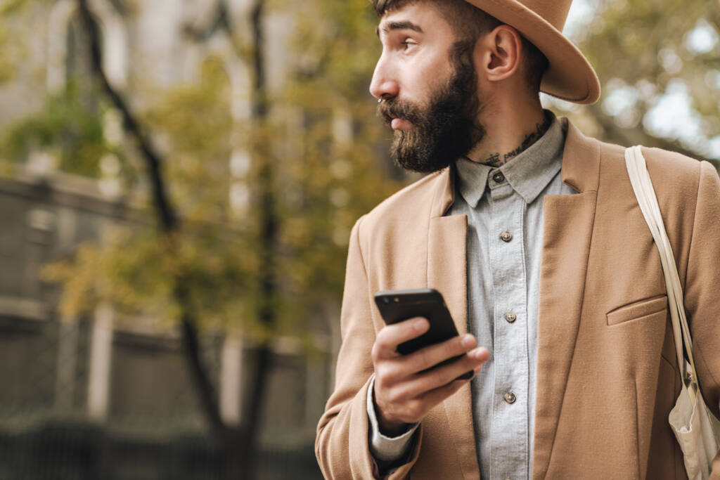 Образ привабливого стильного молодого чоловіка в капелюсі, який тримає мобільний телефон під час прогулянки на відкритому повітрі
 - Фото, зображення