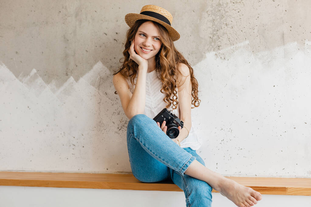 fiatal csinos mosolygós boldog nő visel kék farmer és fehér ing ül a falnak szalma kalap gazdaság vintage fényképezőgép, utazó nyári ruhában, divat trend - Fotó, kép